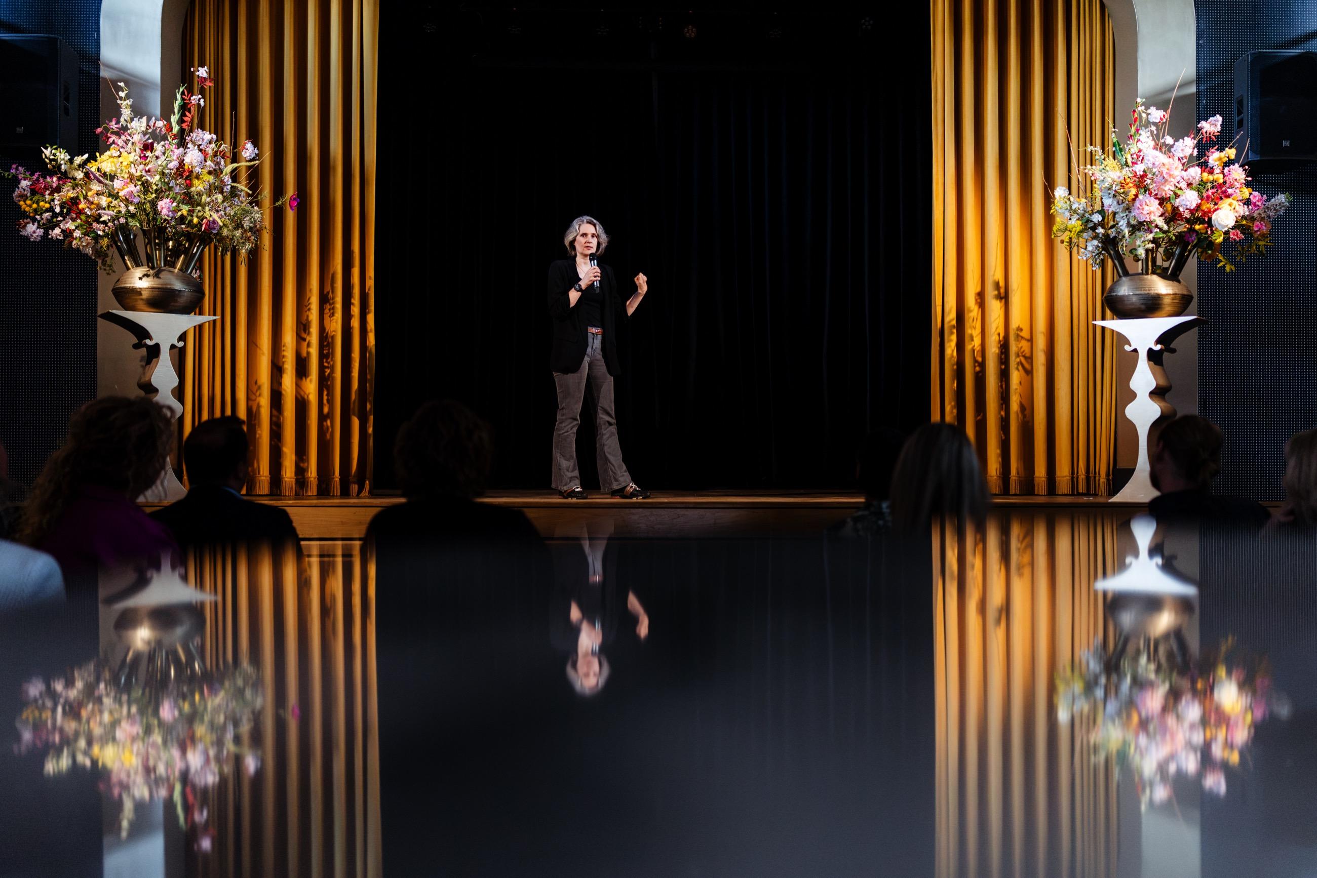 Een spreker staat alleen op een verlicht podium tussen gouden coulissen, publiek op de voorgrond